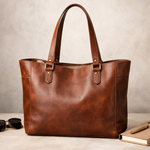 Brown leather tote bag on a neutral background with sunglasses and a book.